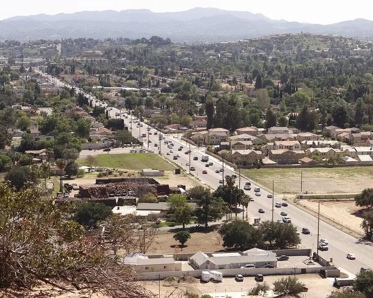A wide boulevard running straight across a suburb toward hazy mountains, seen from a rocky hill